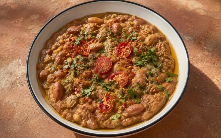 Ful medames in a shallow wide bowl showing thick, rustic partially mashed fava beans in spiced tomato base with olive oil drizzle, fresh parsley, chili flakes, and halved cherry tomatoes on marble surface