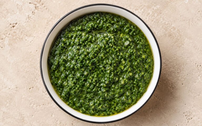 Green zhoug in a small white bowl showing vivid deep green colour with slightly coarse herb texture and olive oil sheen on marble surface with pita bread beside it