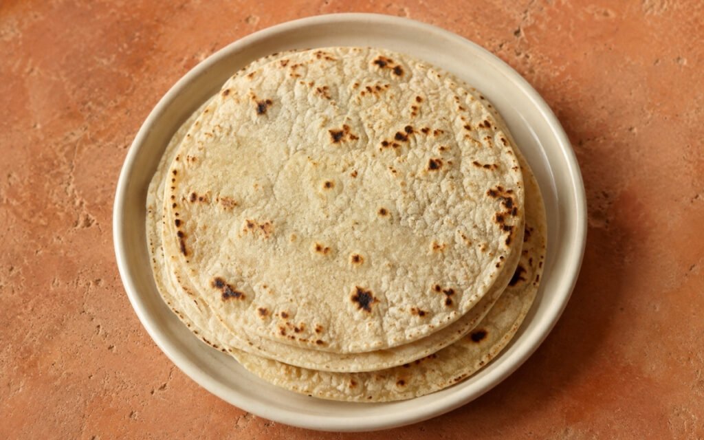 Stack of homemade corn tortillas showing soft texture with irregular char spots on a cloth-lined plate with masa harina and lime wedge beside it
