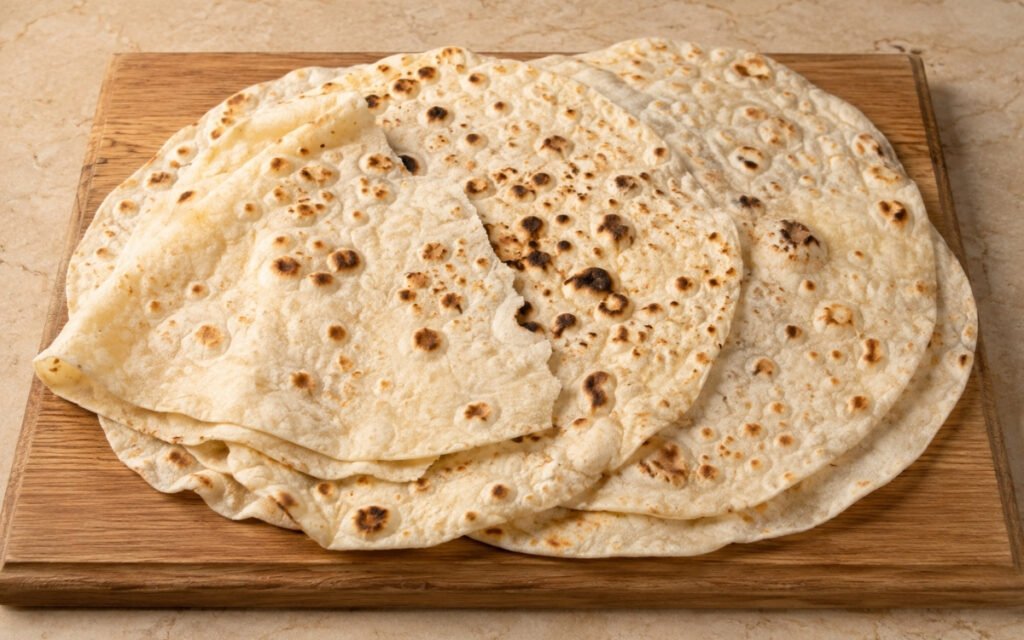 Stack of homemade lavash flatbread showing golden blister spots and soft pliable texture on a cloth-lined wooden board with fresh herbs and dipping sauce beside it