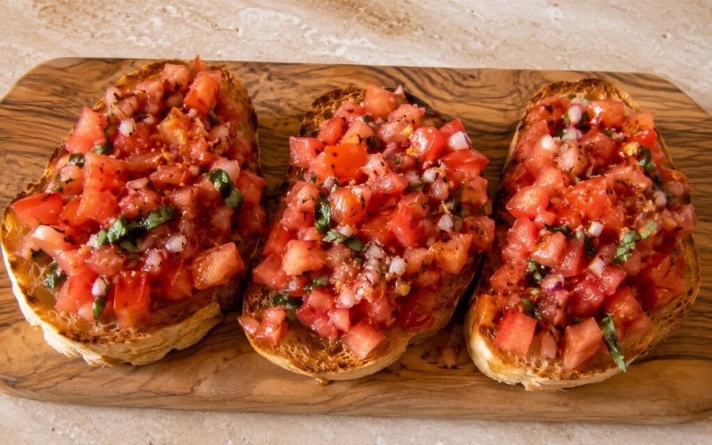 Italian bruschetta al pomodoro on a wooden board showing thick toasted sourdough slices topped with vibrant red heirloom tomato mixture, fresh basil, balsamic drops, and olive oil on marble surface