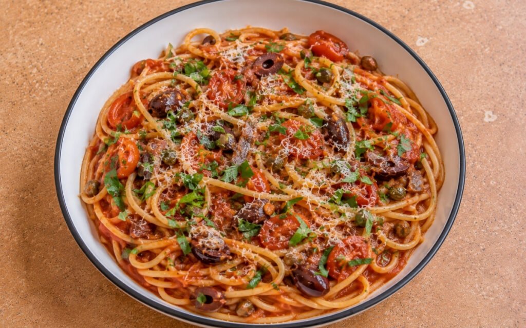 Italian puttanesca pasta with spaghetti in glossy red tomato sauce, halved cherry tomatoes, Kalamata olives, capers, anchovy pieces, fresh oregano and parsley in a white ceramic bowl with black rim