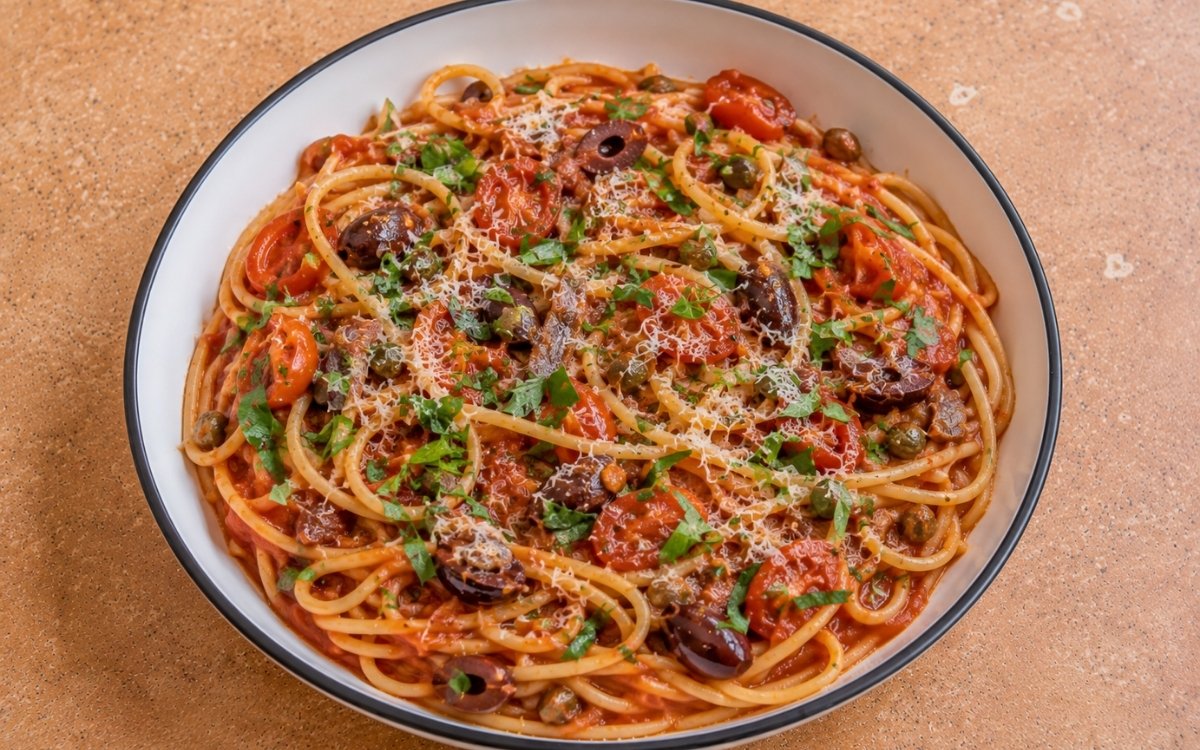 Italian puttanesca pasta with spaghetti in glossy red tomato sauce, halved cherry tomatoes, Kalamata olives, capers, anchovy pieces, fresh oregano and parsley in a white ceramic bowl with black rim