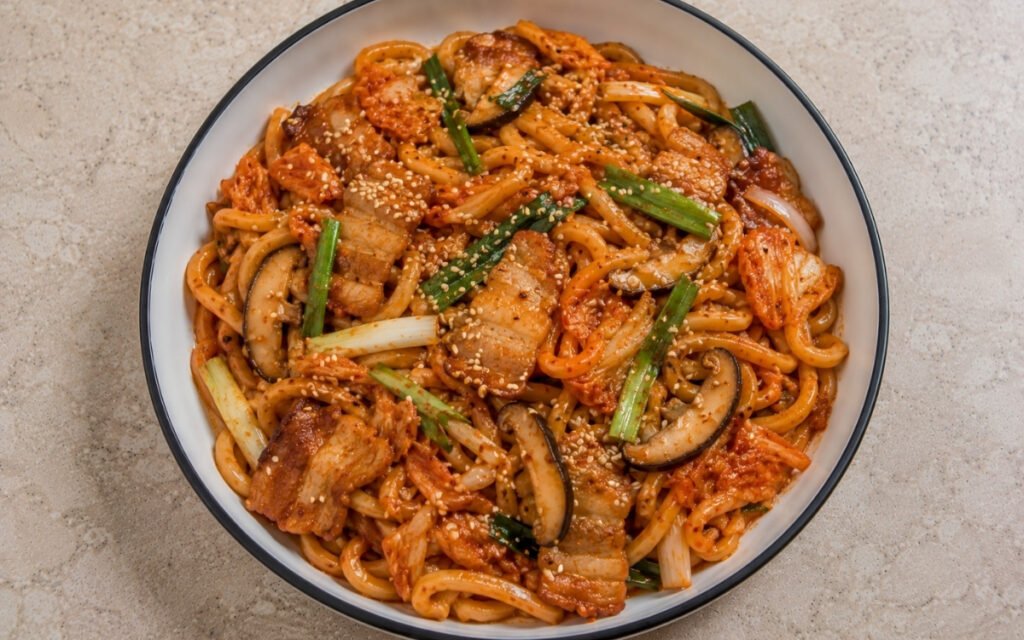 Kimchi udon stir-fry with fresh udon noodles in vivid red-orange kimchi butter sauce, pork belly, chopped kimchi, shiitake, yellow onion, scallions and sesame seeds in a white ceramic bowl with black rim