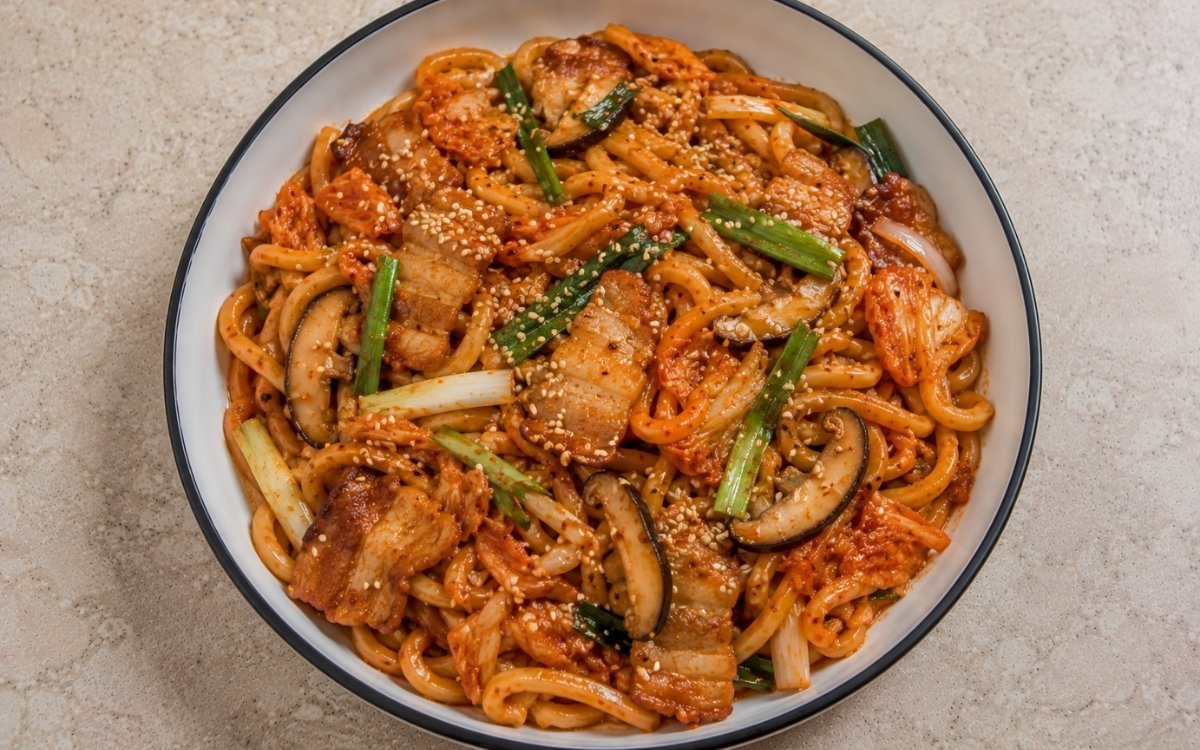 Kimchi udon stir-fry with fresh udon noodles in vivid red-orange kimchi butter sauce, pork belly, chopped kimchi, shiitake, yellow onion, scallions and sesame seeds in a white ceramic bowl with black rim