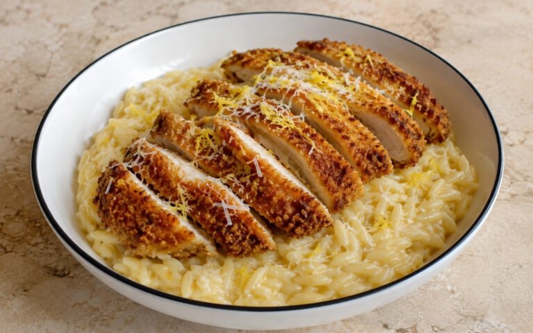 Lemon chicken cutlet orzo in a wide white bowl showing creamy yellow-tinted orzo with sliced golden crispy cutlet strips fanned over the top, Parmesan, and lemon zest on marble surface