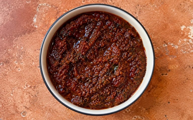 Red zhoug in a small white bowl showing deep brick-red colour with visible cilantro flecks and olive oil sheen on marble surface with pita bread beside it