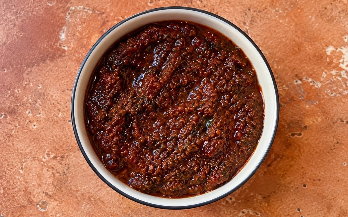 Red zhoug in a small white bowl showing deep brick-red colour with visible cilantro flecks and olive oil sheen on marble surface with pita bread beside it