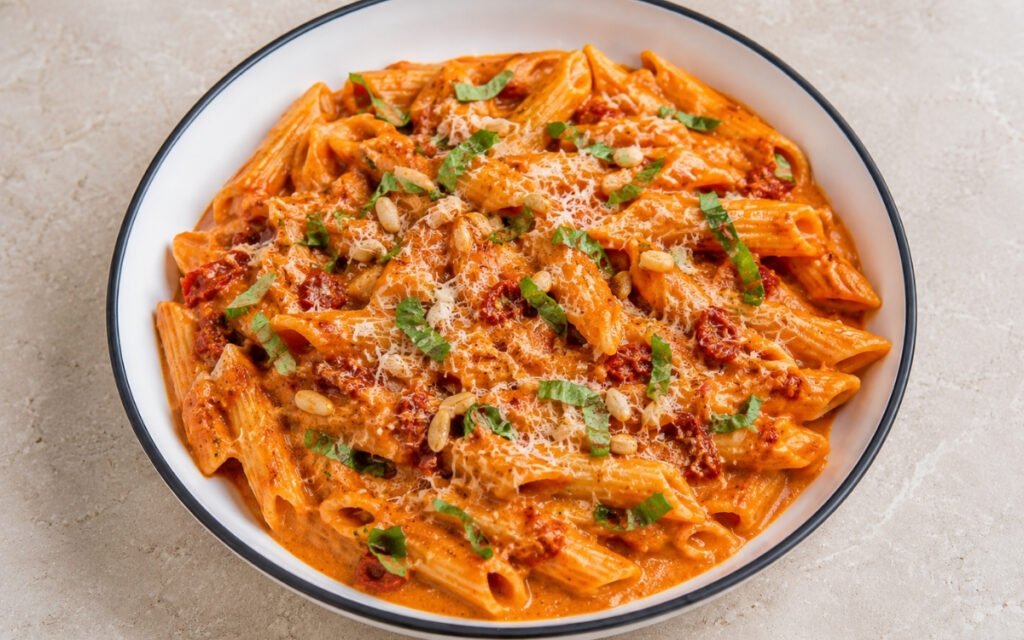 Roasted red pepper cream pasta with penne in glossy vibrant red-orange cream sauce with sun-dried tomatoes, torn basil, toasted pine nuts and Parmigiano-Reggiano in a white ceramic bowl with black rim