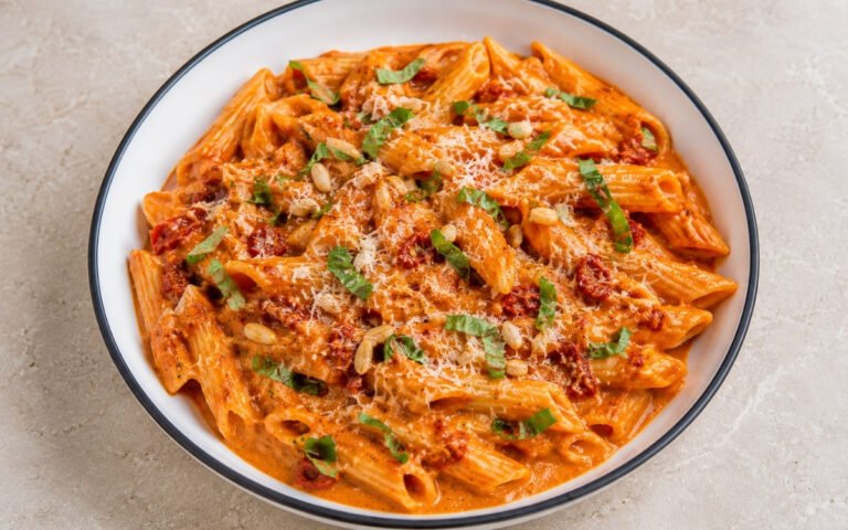 Roasted red pepper cream pasta with penne in glossy vibrant red-orange cream sauce with sun-dried tomatoes, torn basil, toasted pine nuts and Parmigiano-Reggiano in a white ceramic bowl with black rim