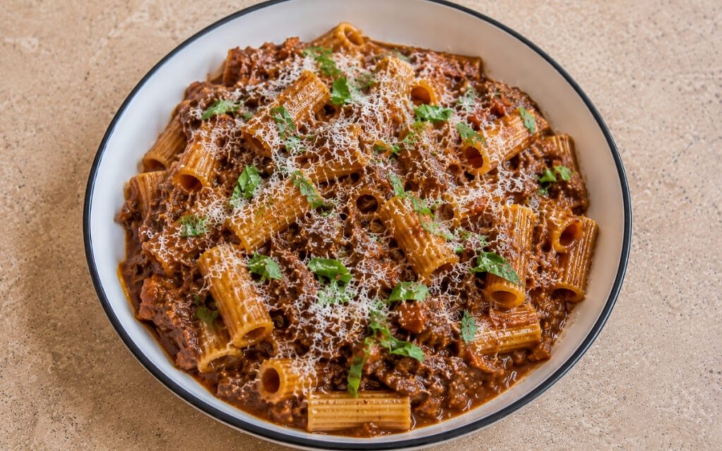 Beef ragù pasta with rigatoni in rich mahogany-brown beef and pancetta ragù with grated Parmigiano-Reggiano and torn fresh basil in a white ceramic bowl with black rim