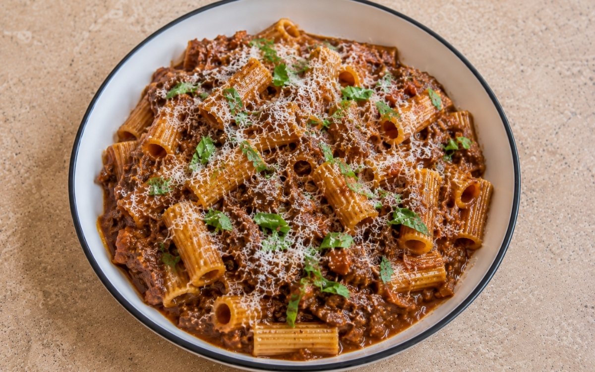 Beef ragù pasta with rigatoni in rich mahogany-brown beef and pancetta ragù with grated Parmigiano-Reggiano and torn fresh basil in a white ceramic bowl with black rim