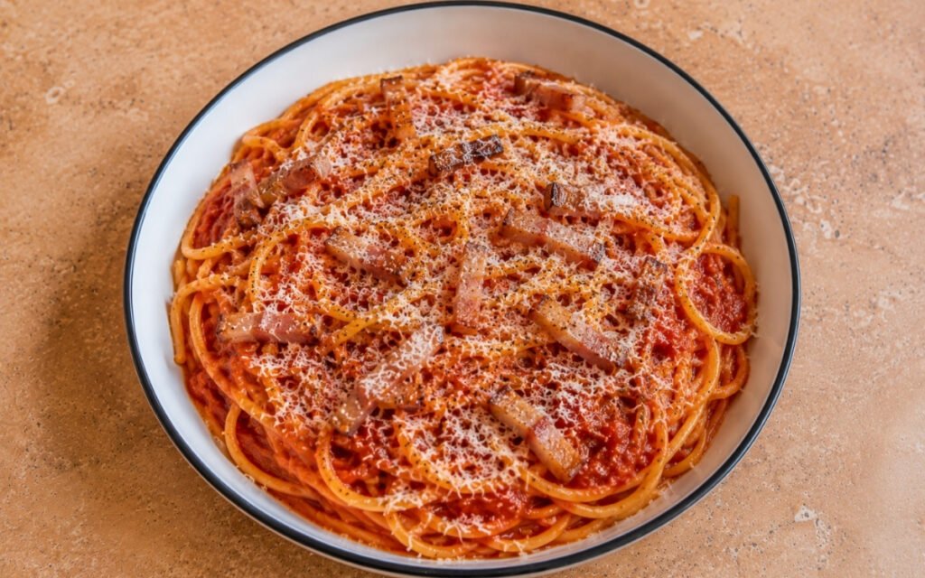 Spaghetti all'amatriciana with bucatini in glossy red San Marzano tomato sauce, crispy golden guanciale strips, grated Pecorino Romano and black pepper in a white ceramic bowl with black rim
