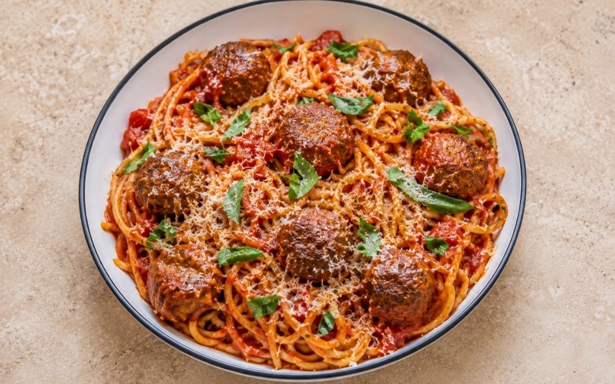 Spaghetti and meatballs in vivid red San Marzano tomato sauce with golden-brown seared meatballs, Parmigiano-Reggiano and fresh basil leaves in a white ceramic bowl with black rim
