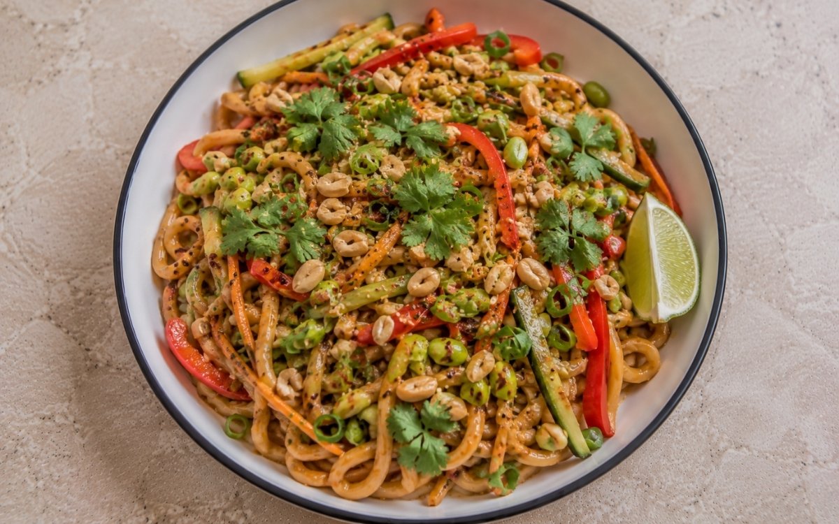 Spicy peanut butter noodles with udon in amber peanut sauce, julienned red bell pepper, carrots, edamame, cucumber, crushed peanuts, cilantro, green onions, chili oil and black sesame seeds in a white ceramic bowl with black rim