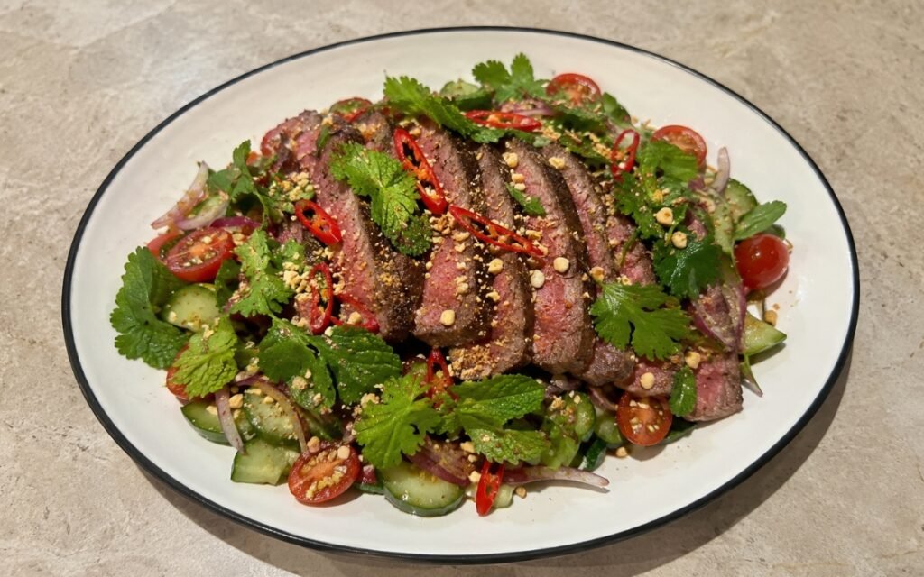 Spicy Thai beef salad on a wide plate showing thinly sliced medium-rare sirloin over fresh mint, cilantro, and Thai basil with cucumber, cherry tomatoes, red onion, and toasted rice powder with visible chili and peanut garnish on marble surface