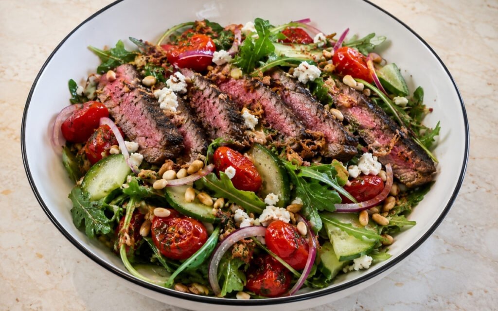 Steak salad bowl with sliced sirloin over mixed greens and arugula with roasted cherry tomatoes, cucumber, red onion, feta, crispy fried shallots, toasted pine nuts and garlic vinaigrette in a white ceramic bowl with black rim