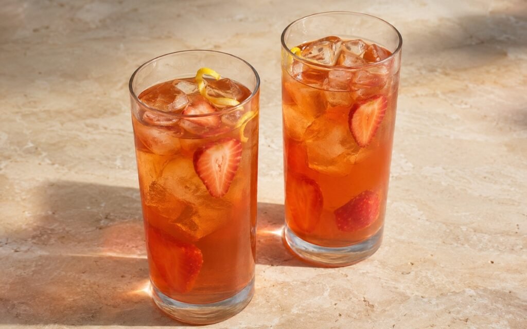 Strawberry iced black tea in a tall glass showing pale pink-amber still drink over ice with fresh strawberry slices on marble surface