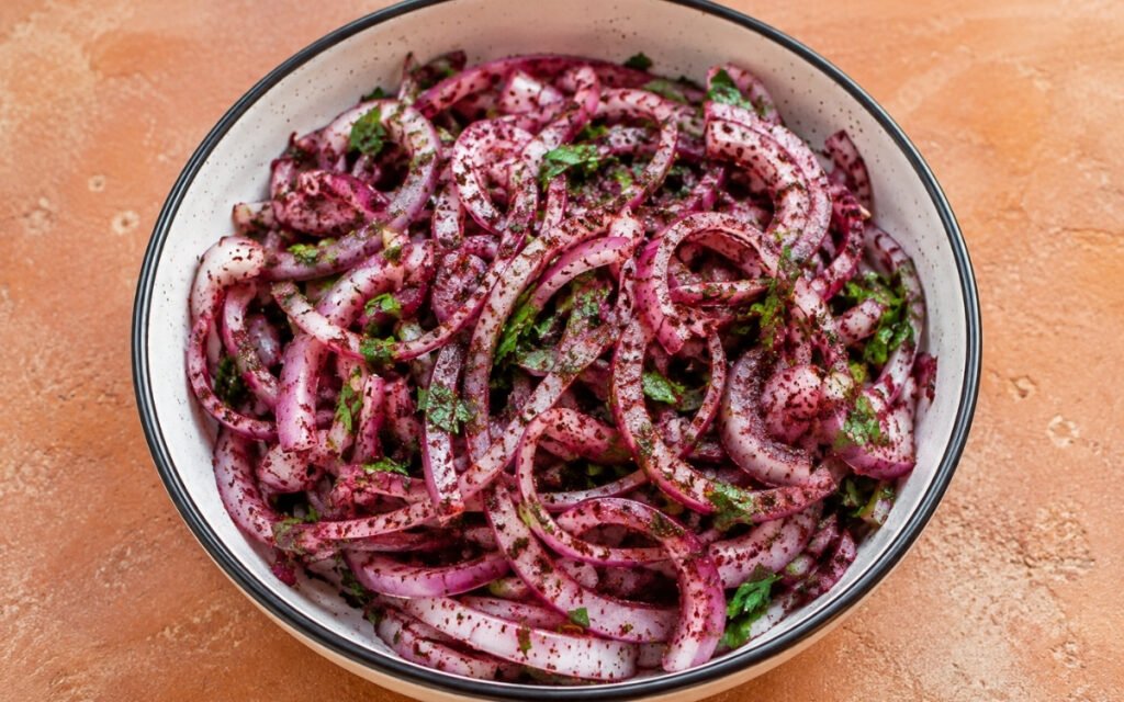 Sumac onions salad in a white bowl showing deep red-purple coated thin onion rings with fresh parsley and visible sumac coating on marble surface