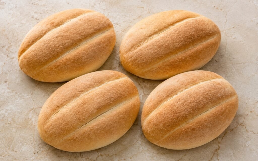 Four golden telera rolls showing deep signature lengthwise grooves and a thin, lightly crisp crust on marble surface