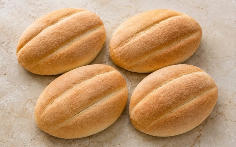 Four golden telera rolls showing deep signature lengthwise grooves and a thin, lightly crisp crust on marble surface
