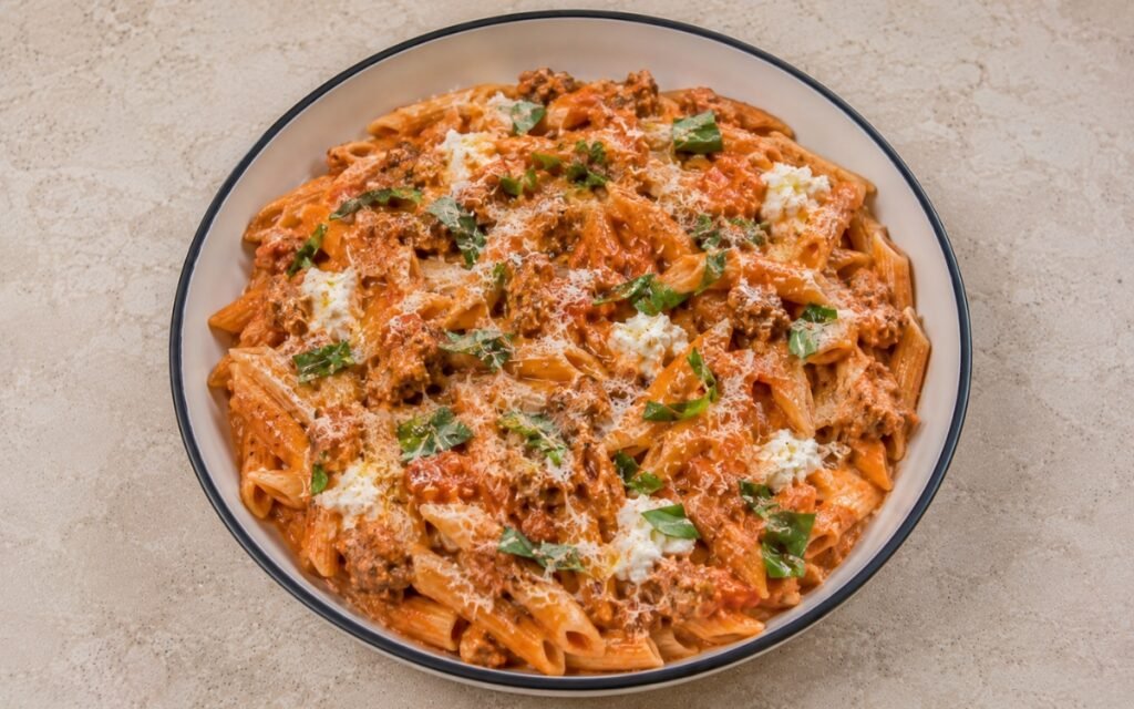 Tomato pasta with ricotta and Italian sausage with fusilli in silky pale pink-red tomato-ricotta sauce, crumbled browned Italian sausage, torn basil and Parmesan in a white ceramic bowl with black rim