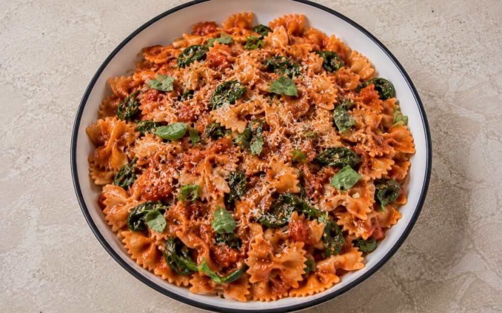 Tomato spinach farfalle pasta with farfalle in glossy vivid red tomato sauce, wilted baby spinach, torn fresh basil, Parmesan and red pepper flakes in a white ceramic bowl with black rim