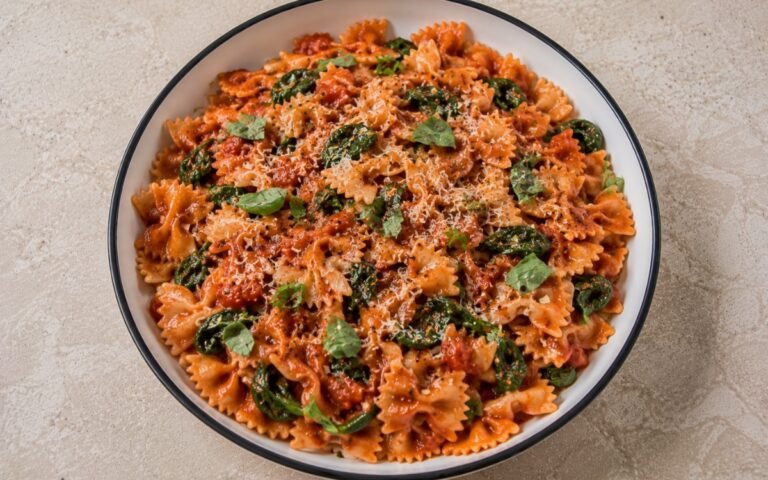 Tomato spinach farfalle pasta with farfalle in glossy vivid red tomato sauce, wilted baby spinach, torn fresh basil, Parmesan and red pepper flakes in a white ceramic bowl with black rim
