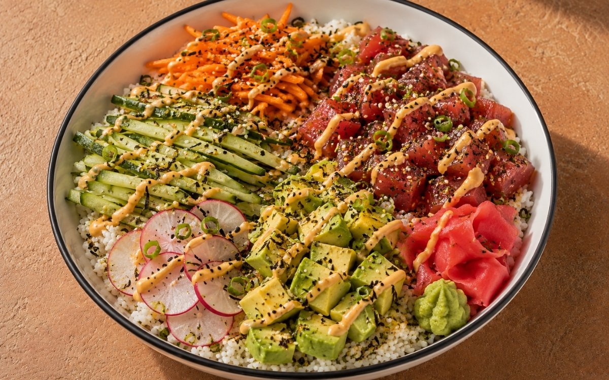 Tuna poke bowl with marinated raw ahi tuna cubes over sushi rice, cucumber, radishes, avocado, carrots, scallions, furikake, spicy mayo, pickled ginger and wasabi in a white ceramic bowl with black rim