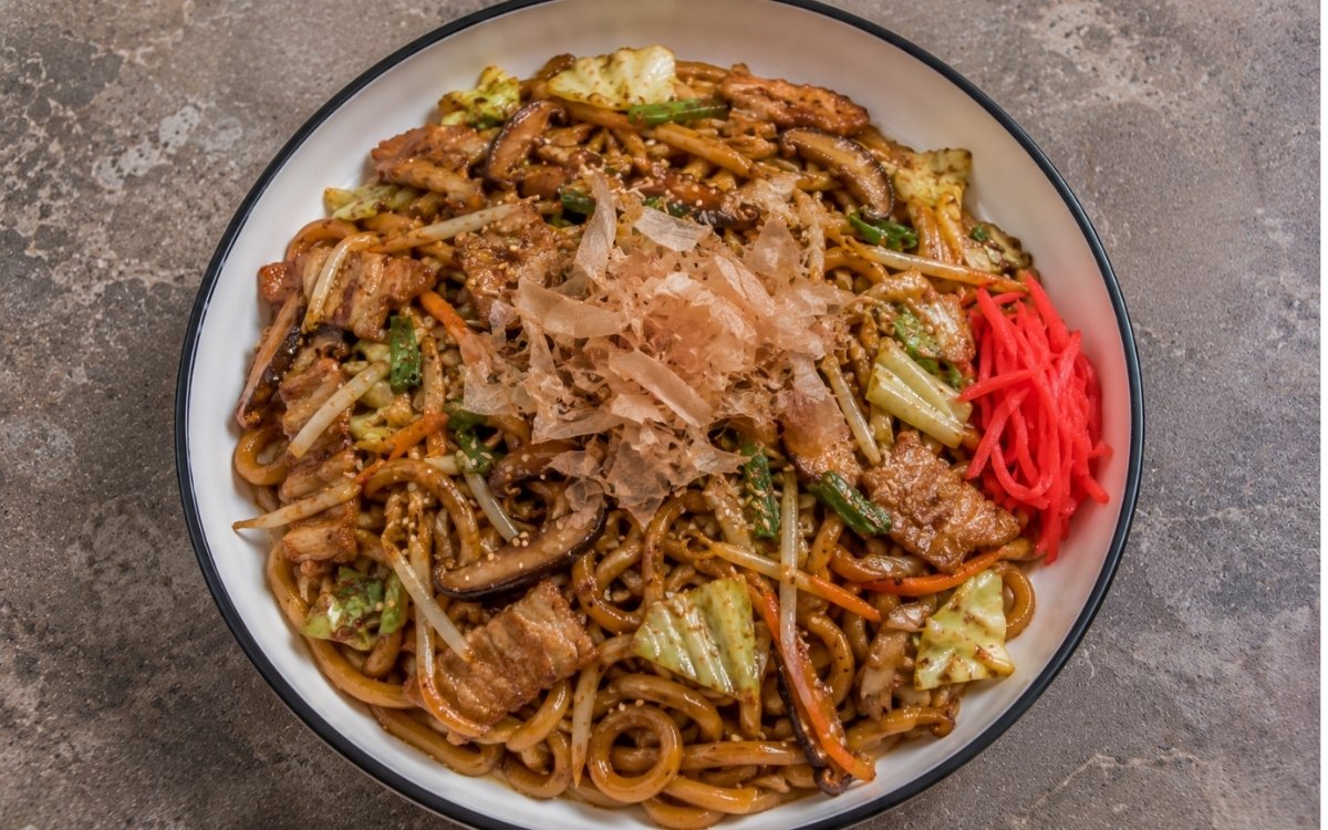 Yaki udon stir-fry with fresh udon noodles in glossy dark soy-mirin sauce, pork belly, shiitake, cabbage, carrots, bean sprouts, scallions, sesame seeds, bonito flakes and pickled red ginger in a white ceramic bowl with black rim