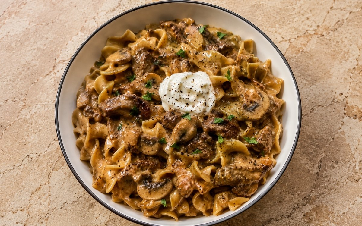 Beef stroganoff showing tender beef strips and mushrooms in a creamy brown sauce over buttery egg noodles, topped with sour cream and parsley, on marble surface