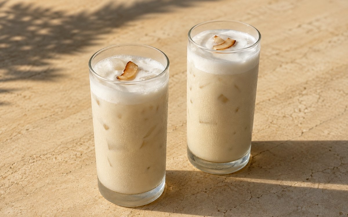 Coconut iced latte in a tall glass showing a very pale coconut-cream beige colour with a soft white coconut cream layer on top and toasted coconut flakes, on marble surface