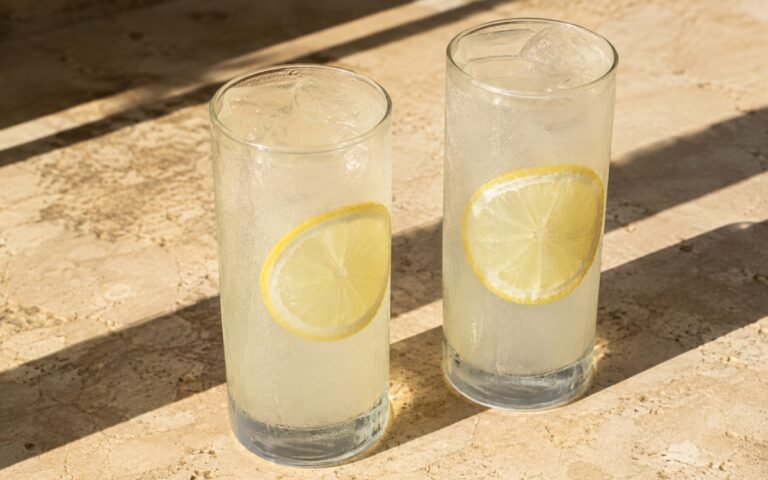 glasses of pale, nearly-clear coconut lemonade with lemon wheels floating in ice, on marble surface