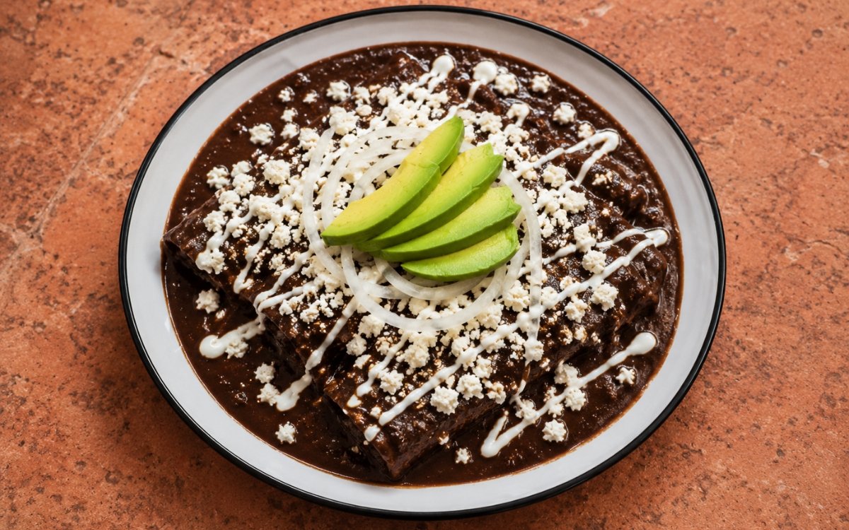 Enfrijoladas showing folded corn tortillas coated in glossy black bean sauce, topped with crumbled queso fresco, crema, thin onion, and sliced avocado, on marble surface