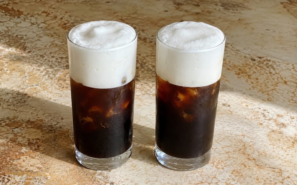 Freddo Cappuccino in a tall glass showing dark chilled espresso over ice topped with a thick, dense white milk foam cap, on marble surface