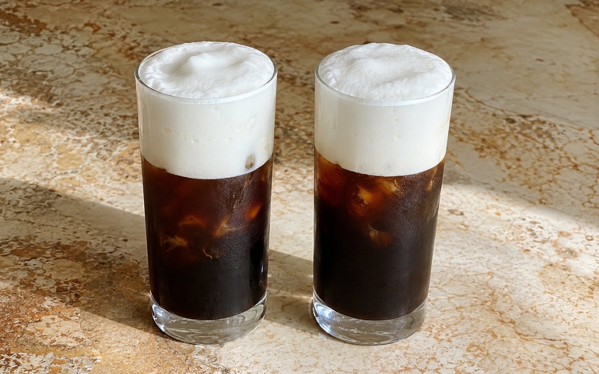 Freddo Cappuccino in a tall glass showing dark chilled espresso over ice topped with a thick, dense white milk foam cap, on marble surface