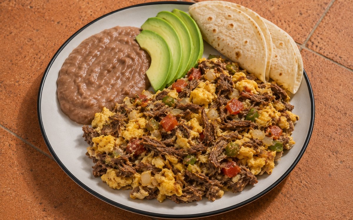 Plate of machaca con huevo showing scrambled eggs with shredded dried beef, tomato, and onion, served with refried beans, sliced avocado, and warm flour tortillas, on marble surface