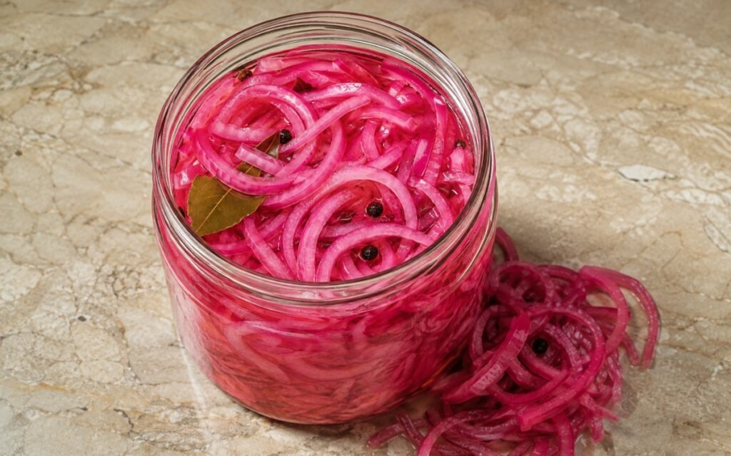 Jar of vivid pink pickled red onion showing crisp-tender slices submerged in brine with visible bay leaf and peppercorns, on marble surface