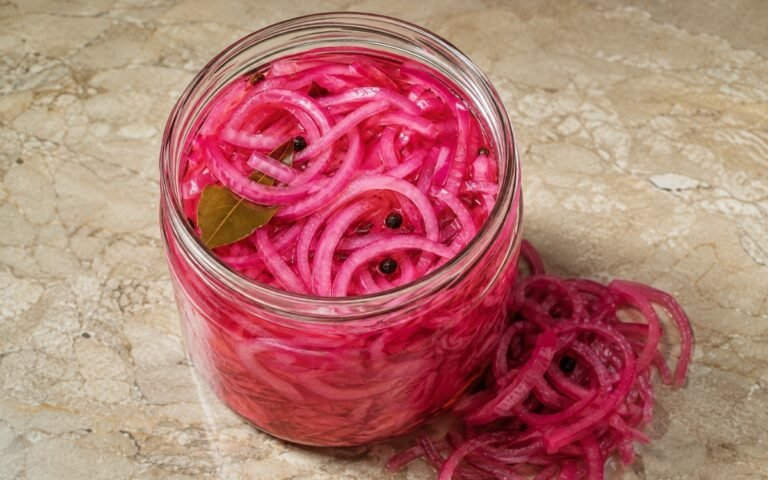 Jar of vivid pink pickled red onion showing crisp-tender slices submerged in brine with visible bay leaf and peppercorns, on marble surface