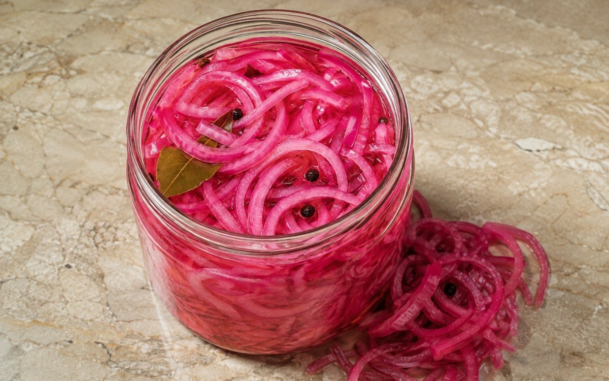 Jar of vivid pink pickled red onion showing crisp-tender slices submerged in brine with visible bay leaf and peppercorns, on marble surface