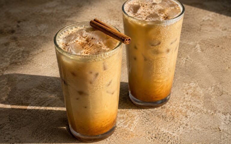 Pumpkin spice iced latte in a tall glass showing a deeper amber-orange base fading into pale caramel-cream milk, dusted with cinnamon and nutmeg, on marble surface