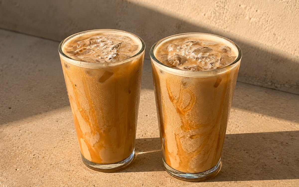 Salted caramel iced latte in a tall glass showing caramel drizzled inside the glass, a rich golden-caramel milk body, and flaky sea salt over a thin foam layer, on marble surface
