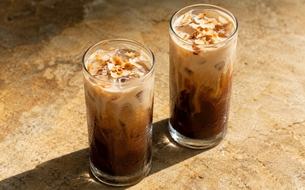 Toasted coconut cold brew in a tall glass showing dark cold brew coffee with soft pale coconut milk swirls moving through it over ice, on marble surface