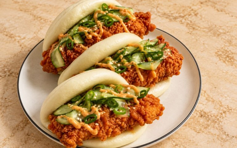 Bao buns showing pillowy steamed buns filled with craggy crisp fried chicken, pickled cucumber ribbons, and glossy sriracha mayo, on marble surface
