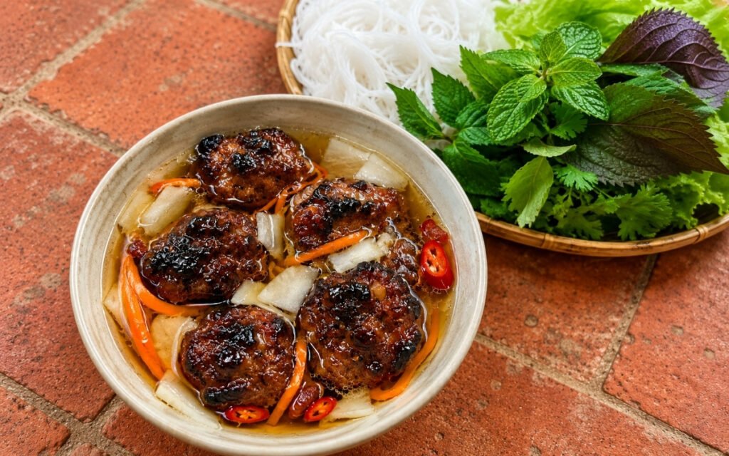 Bún chả showing charred grilled pork patties in warm nước chấm broth with pickled carrot and daikon, served with cold rice vermicelli and fresh herbs, on marble surface