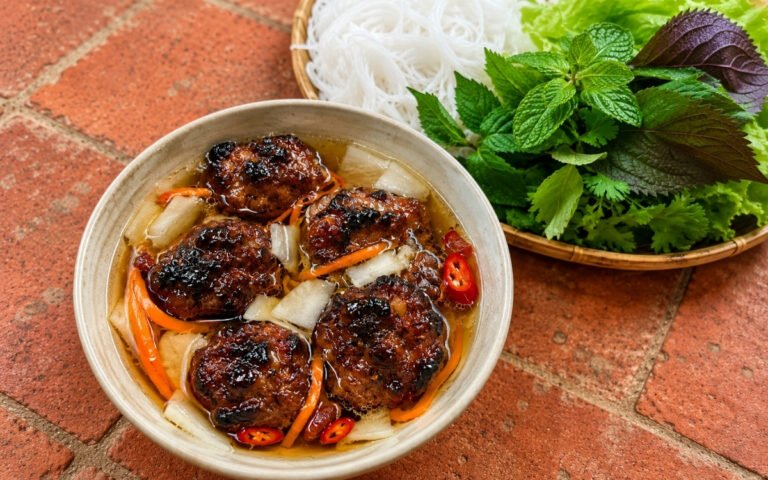Bún chả showing charred grilled pork patties in warm nước chấm broth with pickled carrot and daikon, served with cold rice vermicelli and fresh herbs, on marble surface
