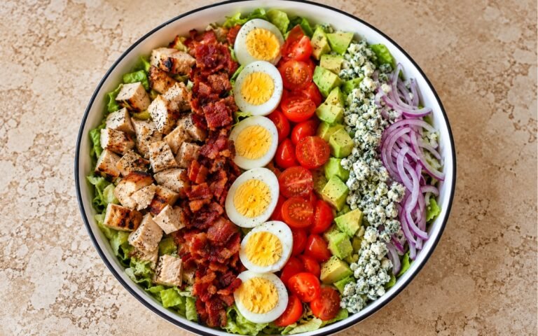 Classic Cobb salad showing neat composed rows of chicken, bacon, eggs, tomatoes, avocado, and blue cheese over chopped lettuce, on marble surface