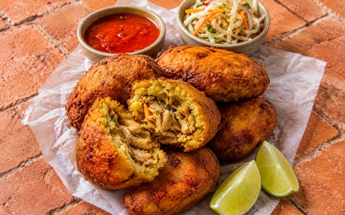 Corviche de pescado showing deeply golden fried plantain dumplings with savory fish filling, served with lime and salsa, on marble surface