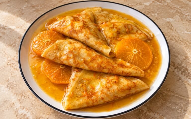 Crêpes Suzette showing folded crêpe triangles coated in glossy orange-butter sauce with fresh orange segments, on marble surface