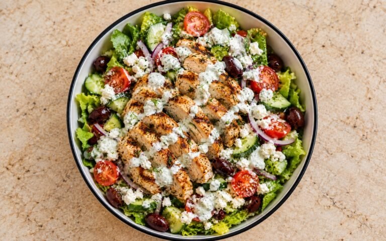 Greek chicken salad showing sliced grilled chicken over crisp greens with cucumber, tomatoes, Kalamata olives, feta, and tzatziki dressing, on marble surface
